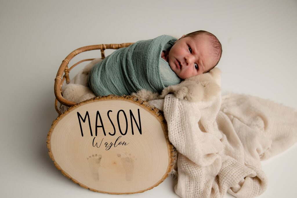 Baby boy laying in basket with sign and blanket in front of it.
