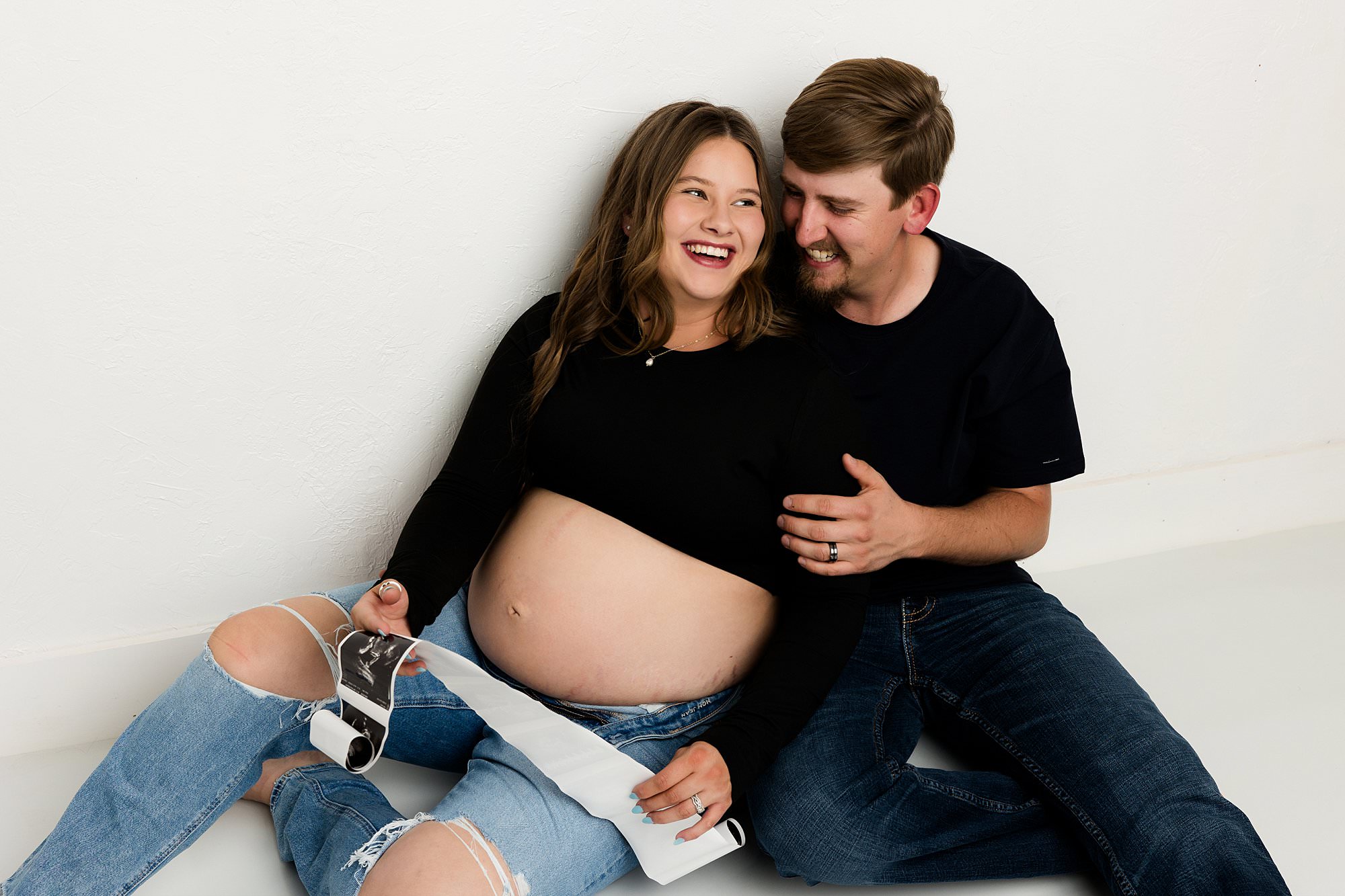 Maternity couple sitting on studio floor with a sonogram paper in mama's hands. Couple is both wearing denim jeans with a black shirt. Studio maternity session photos taken by Lubbock maternity photographer Mary Weibe