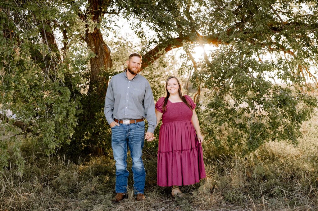 husband and wife standing in front of trees holding hands smiling at camera