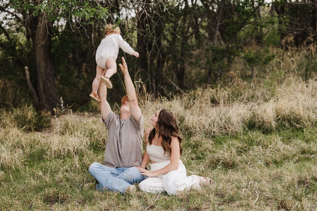 family of three sitting on the grass playing with their little girl, dad is tossing her in the air and catching her. momma is looking at the two of them playing.