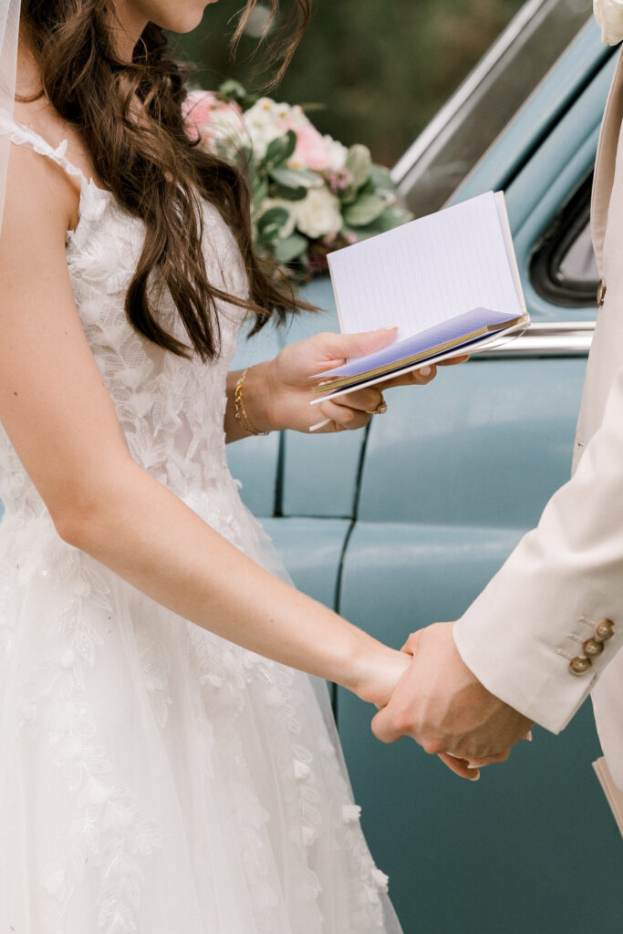 Lubbock wedding photographer captures emotional first look between bride and groom in front of vintage blue car. Bride in white gown with pink rose bouquet smiles at groom in beige suit during romantic Texas wedding. Bride is reading her vows to the groom.