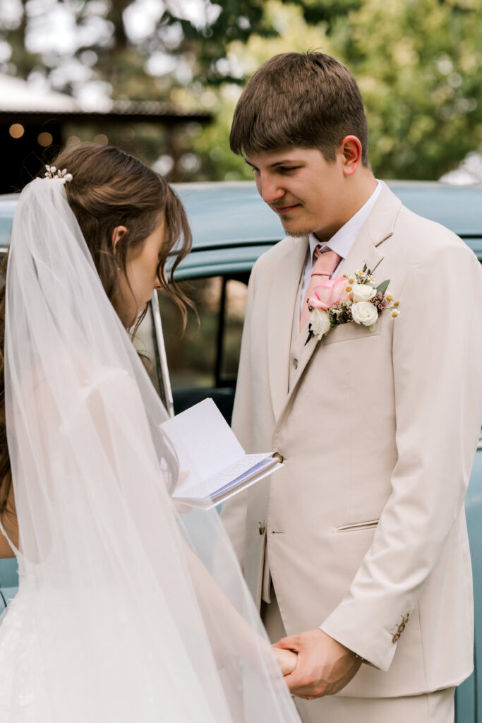 Lubbock wedding photographer captures emotional first look between bride and groom in front of vintage blue car. Bride in white gown with pink rose bouquet smiles at groom in beige suit during romantic Texas wedding