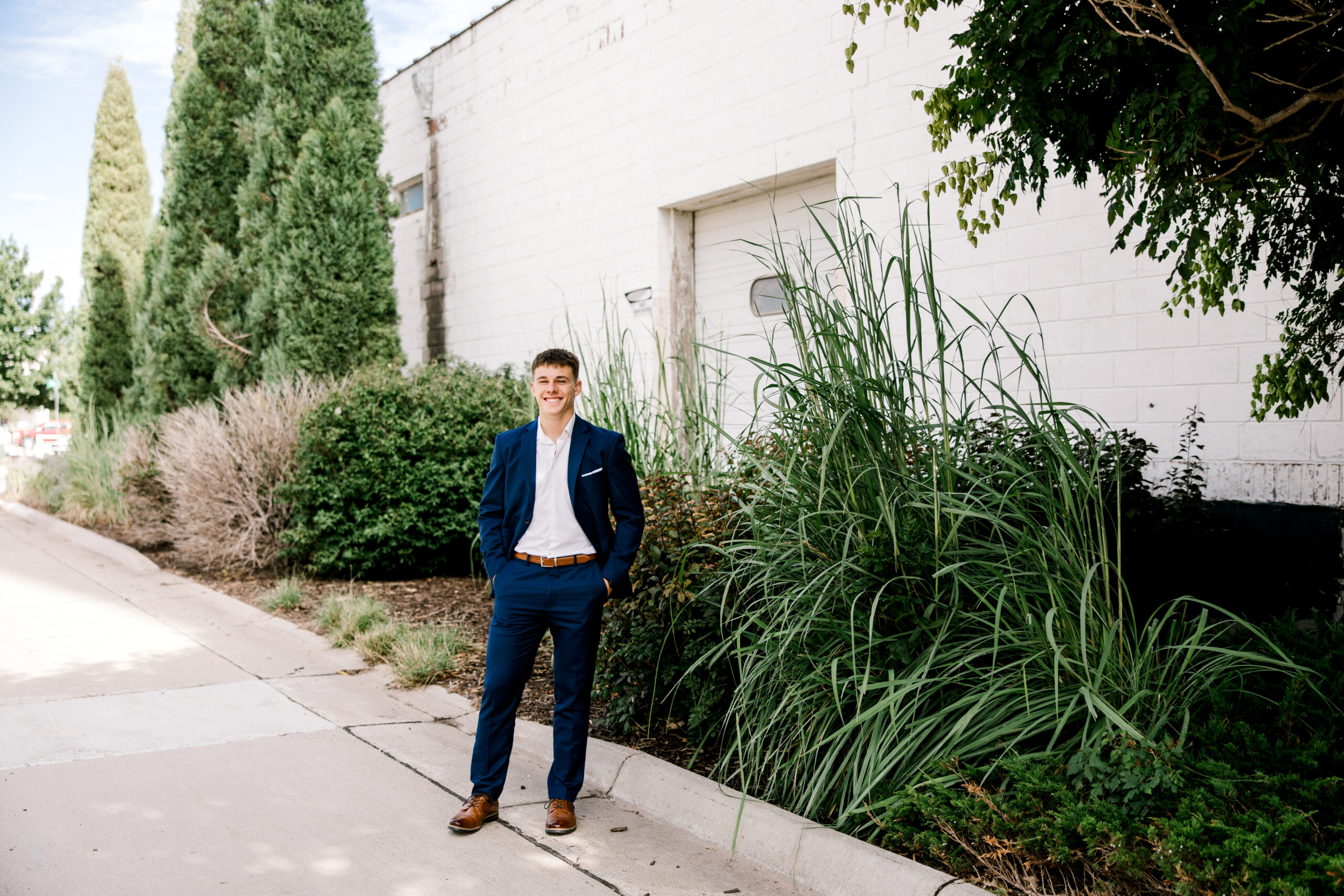 Lubbock Senior photographer Mary Wiebe captures a senior guy dressed in a navy blue suit next to a white building lined with green shrubs and trees. Photo taken in Garden City, Kansas.
