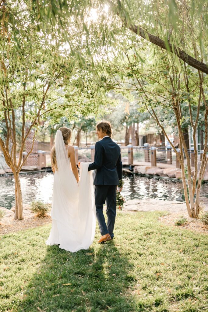Bride and groom walking toward the pond at the wedding venue, groom is holding the train of the bride's dress.