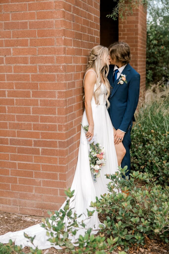 Bride is leaned up against a brick wall, groom leans in to steal a kiss. Wedding at the wedding venue in Lubbock Texas, the Willows