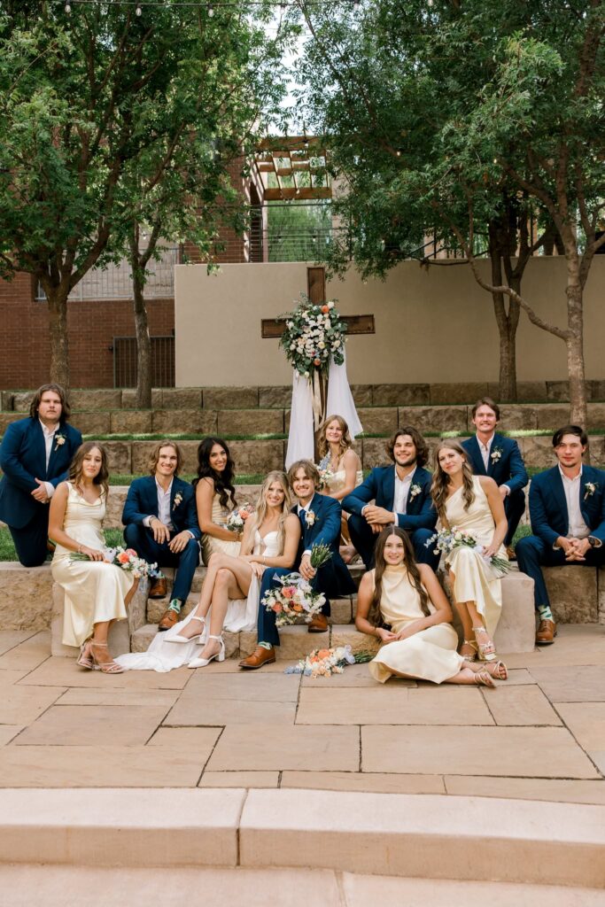 Bridal party portrait. Bridesmaids are in light yellow dresses and groomsmen are wearing navy blue suits. The photo is of the whole bridal party together, they are sitting on the steps of the altar, staggered with a variety of poses.