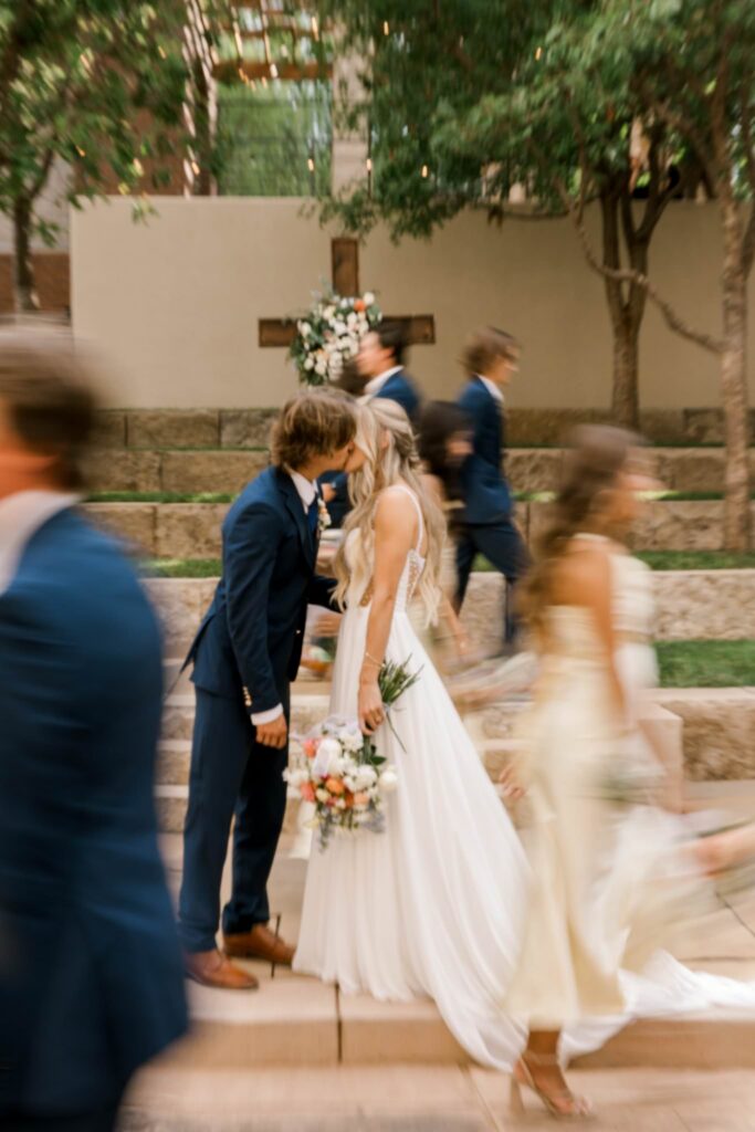Groom leaning in for a kiss as the bridal party passes by them creating a blurry photo effect.