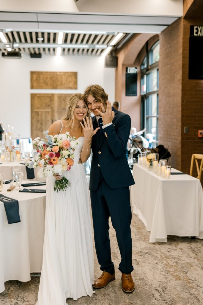 Briana and Colton's wedding day at the Willows Event Center. Bride and groom, smiling at the camera, both are holding up their wedding rings.