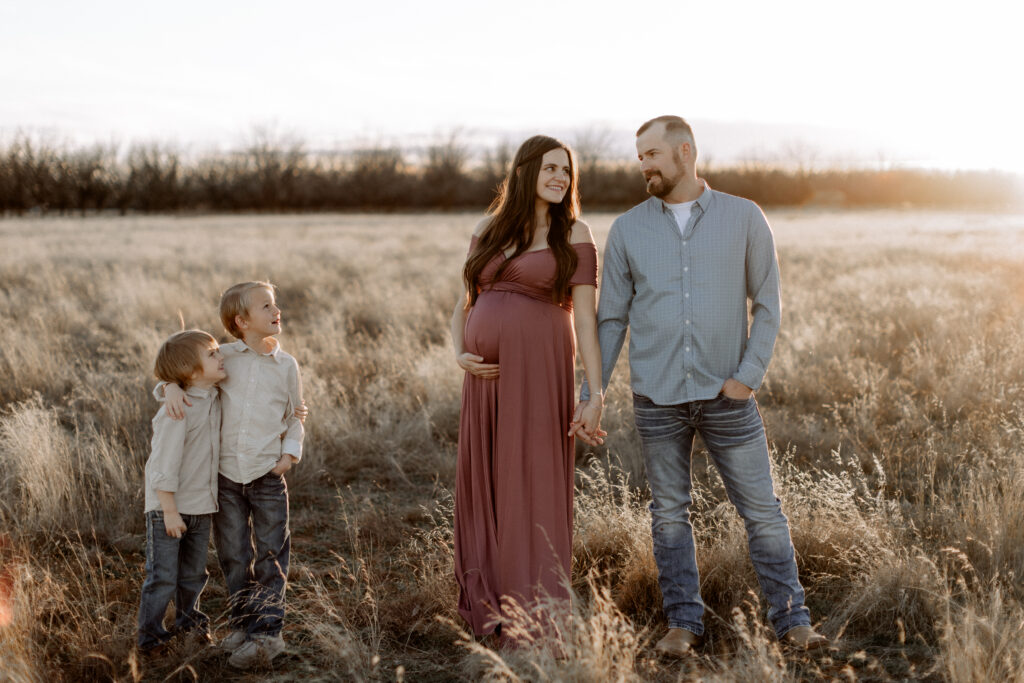 It is worth capturing every pregnancy. In this maternity session the mom and dad are holding hands, and their two little boys are smiling up at them from a few feet away. Captured by Mary Wiebe Photography.