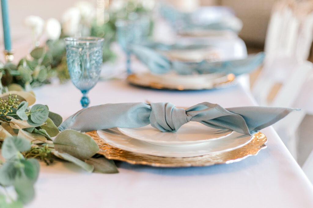 reception day details, elegant white dinner plate, with a light blue napkin tied in a knot, with blue drinking glasses.