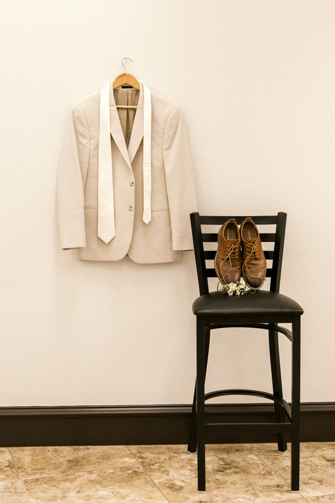 Grooms details, his suit and tie are hung on the wall and his shoes and boutonnière are on a chair next to the suit.