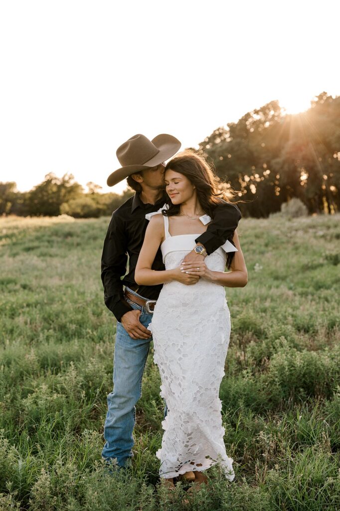 Future bride and groom standing in a green field with trees in the background. Captured by Mary Wiebe Photography. 