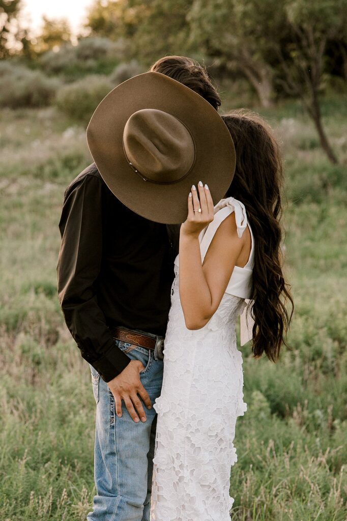 She is holding her future grooms cowboy hat while they steel a kiss hiding behind the hat. 