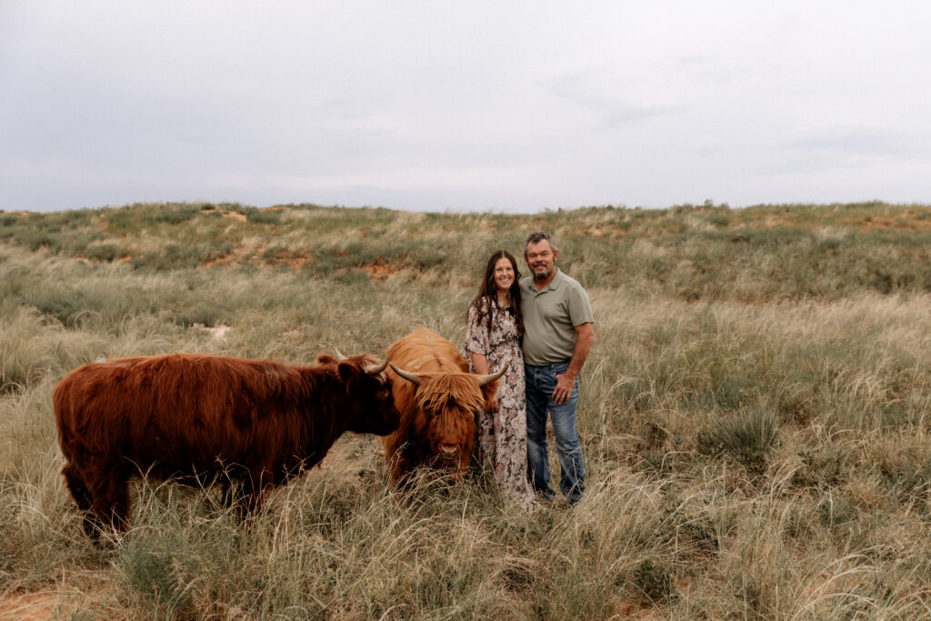 Mary Wiebe and her husband standing in a field with two of her highland cows. The heart behind Mary Wiebe Photography.