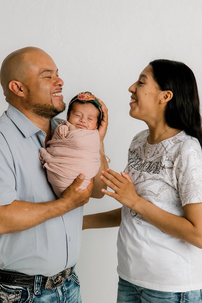 Mom and dad are laughing and smiling while holding their baby girl who is also smiling. 