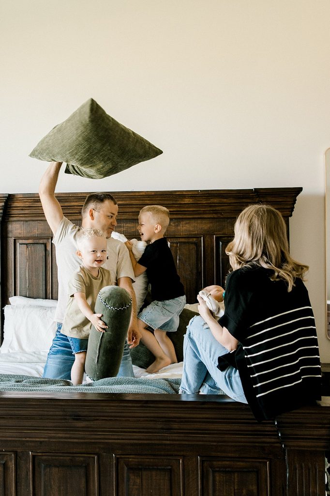 Dad and two older boys having a pillow fight in bed while mama holds her newborn smiling.