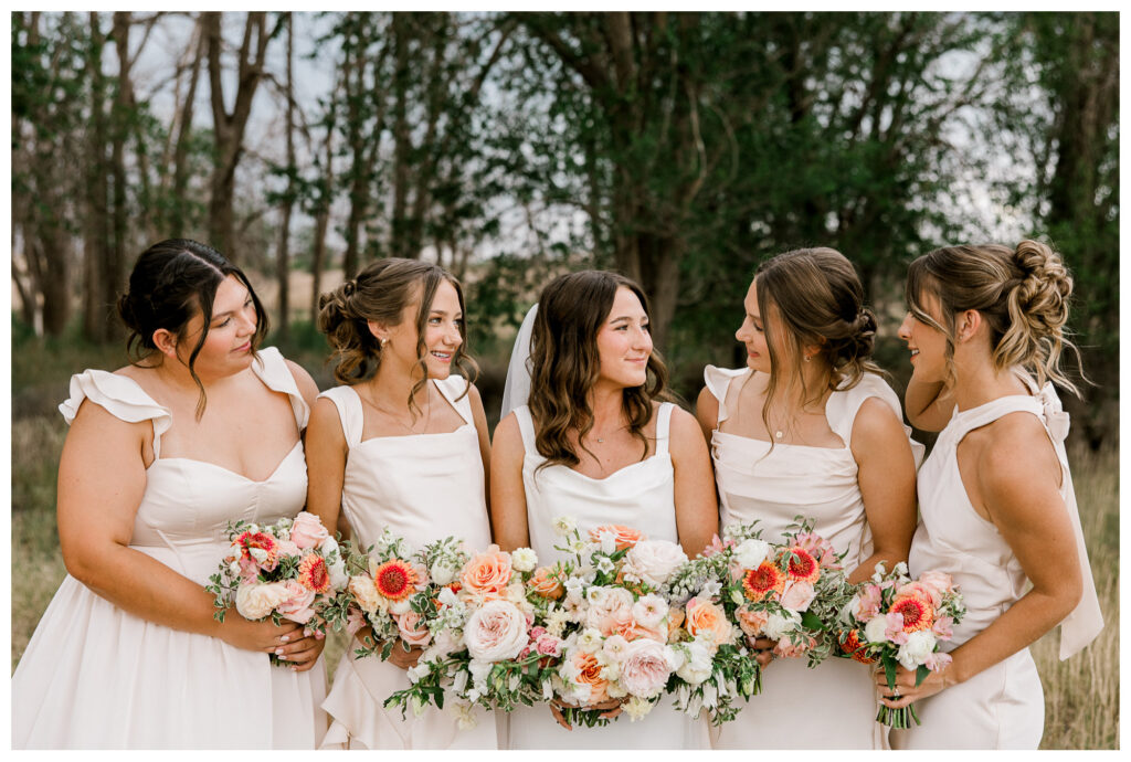 Bridesmaids in ivory dresses holding blush pink, peach, and coral bouquets against a natural woodland background