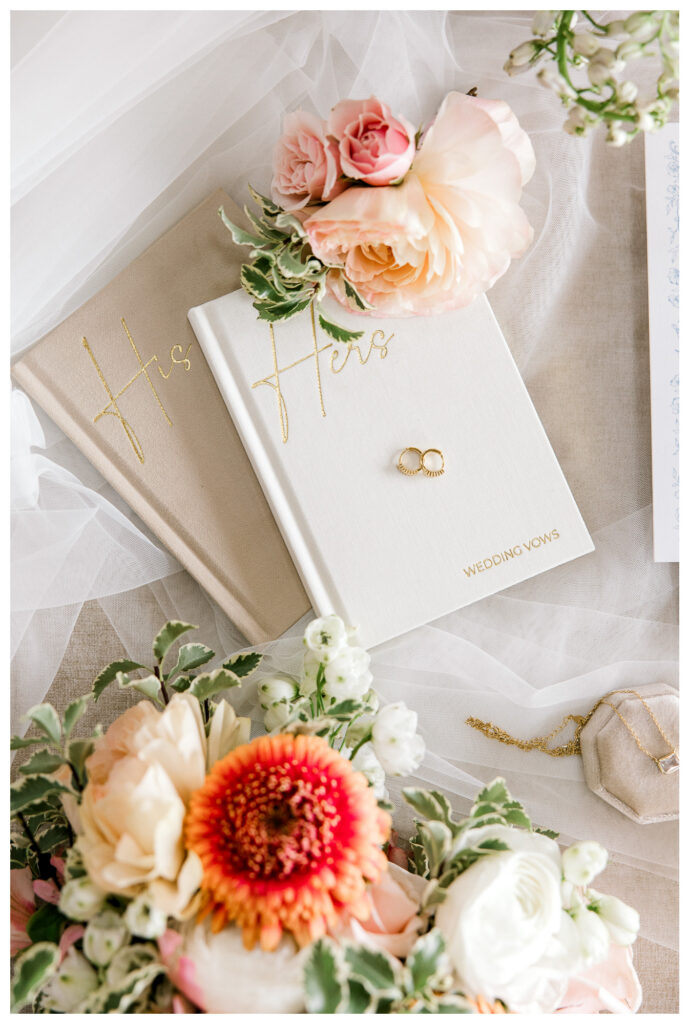Wedding vows books and gold rings styled with ivory fabric, peach roses, and coral flowers in a romantic wedding color palette