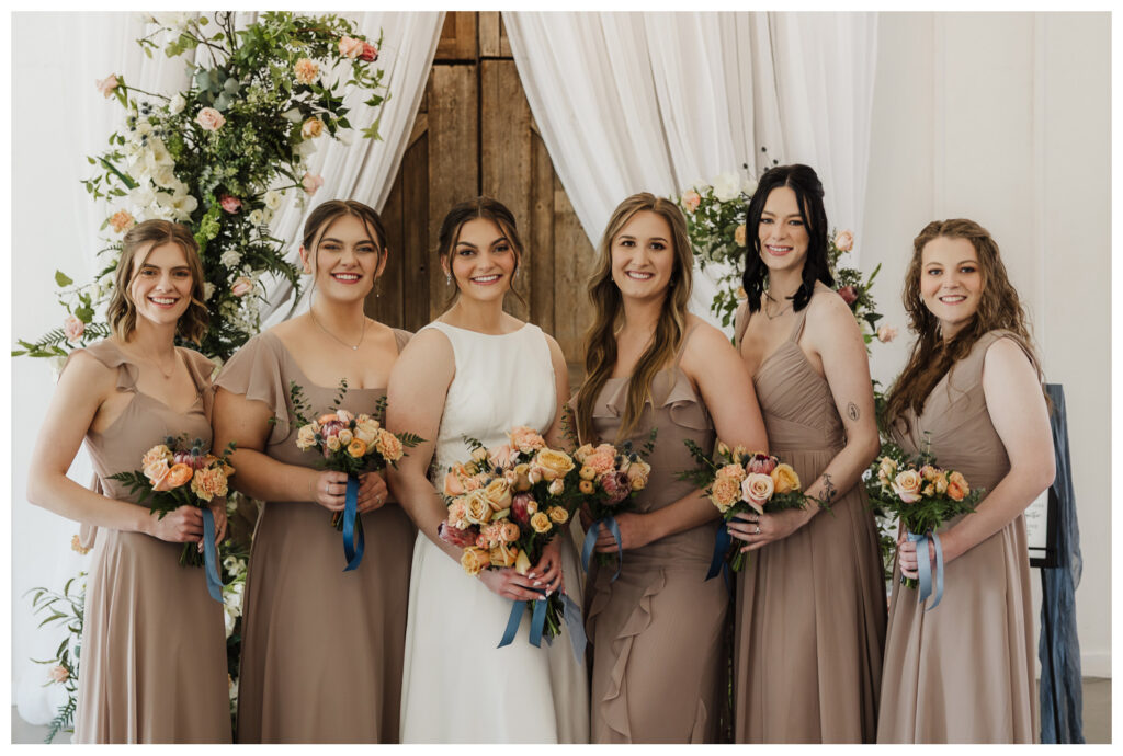 Bridesmaids in taupe and champagne dresses holding peach and blush bouquets with blue ribbon accents